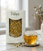 A box of Camille chamomile tea on a wooden kitchen counter beside a window, surrounded by fresh chamomile flowers and a steaming cup of brewed tea in soft natural light.
