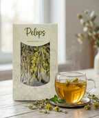 A pack of Pelops Greek mountain tea on a kitchen counter beside a window, surrounded by dried Sideritis stems and a steaming cup of brewed tea in warm natural light.