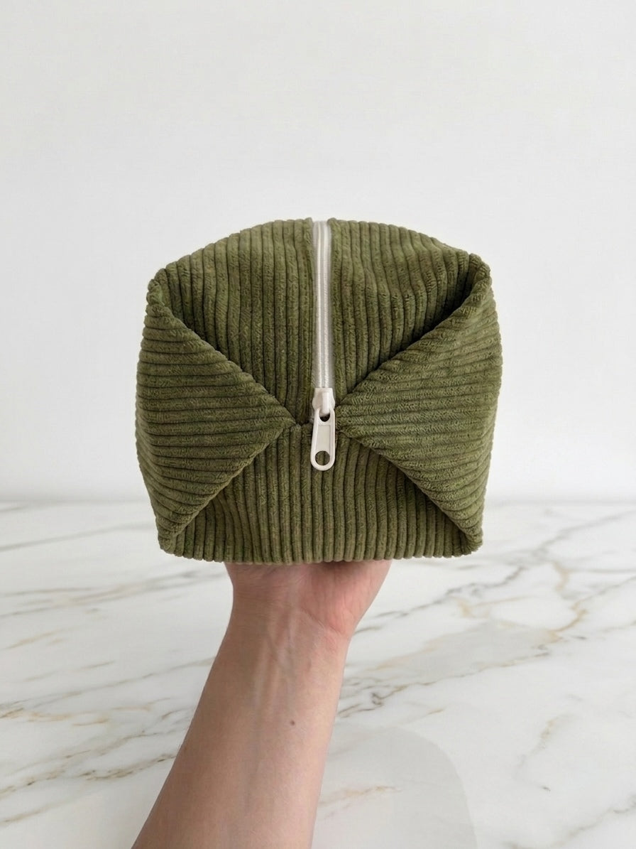 An olive green corduroy zippered pouch held in a hand against a white marble surface and background.