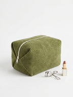 An olive green corduroy makeup bag with a silver zipper, styled on a white background next to a lipstick and an eyelash curler.