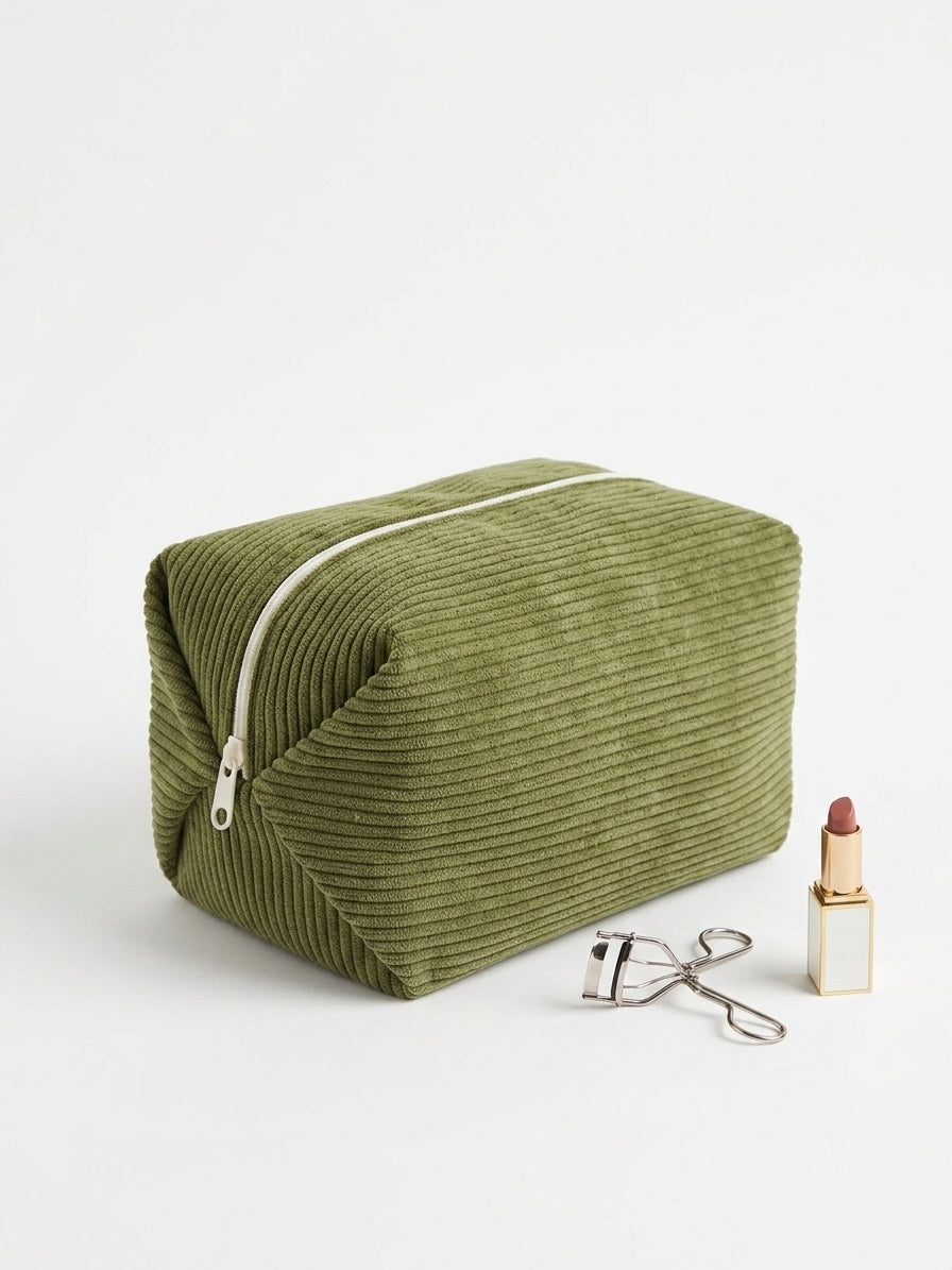 An olive green corduroy makeup bag with a silver zipper, styled on a white background next to a lipstick and an eyelash curler.
