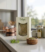 A pack of Pelops Greek mountain oregano on a kitchen counter beside a window, surrounded by dried oregano sprigs and a small dish of olive oil in warm natural light.